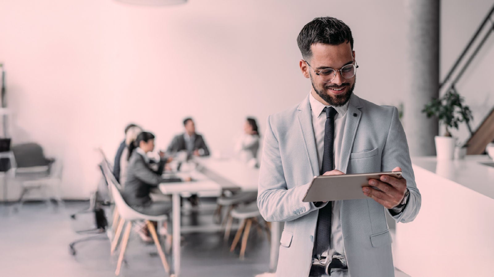 A professional in business attire stands in focus with a tablet, while a group of colleagues collaborate at a conference table in the blurred background.