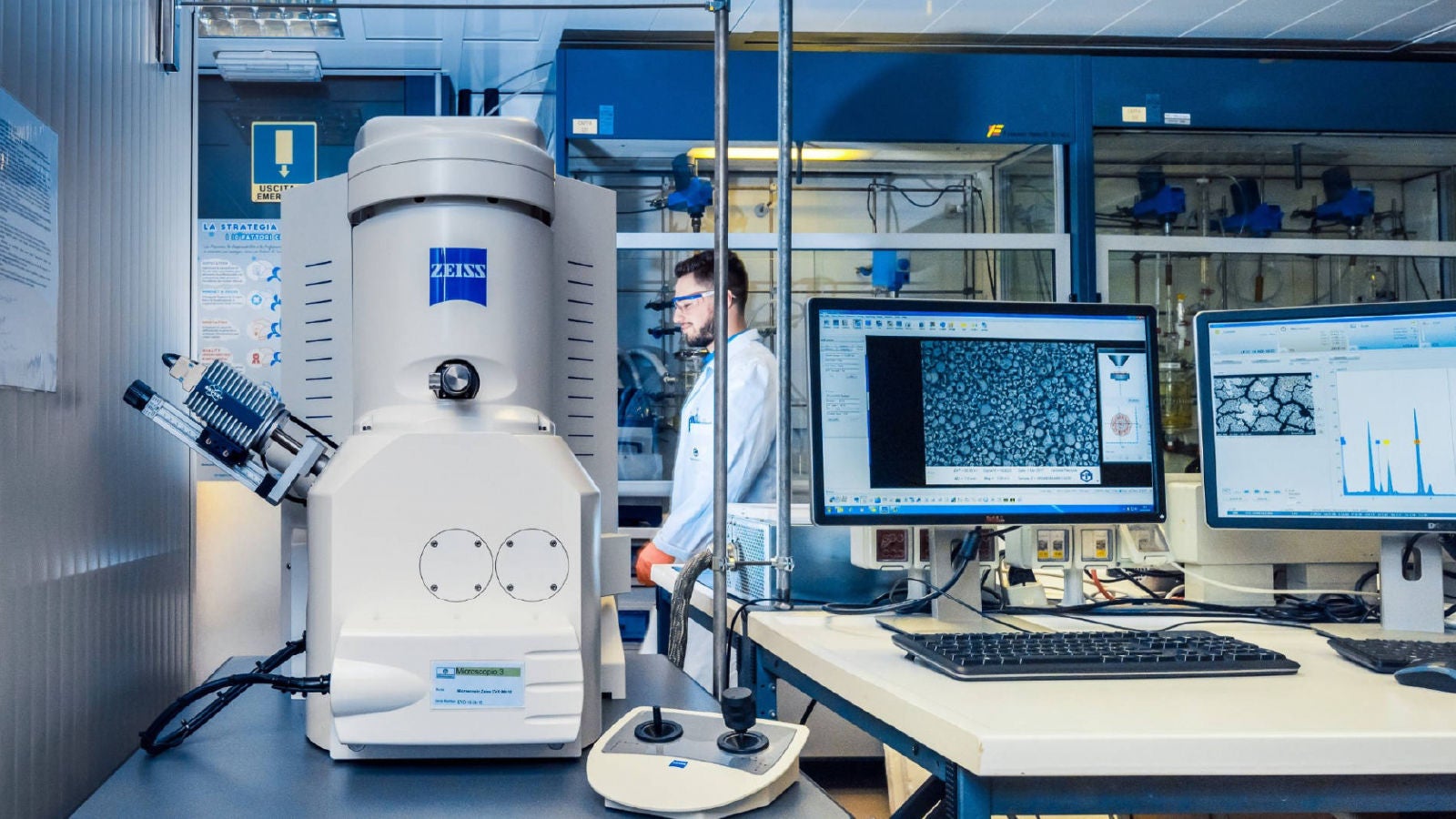 A close-up of laboratory equipment and computer monitors displaying microscopic data and analysis charts, with a scientist working in the background.