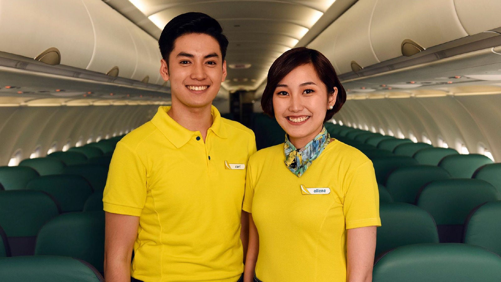 Two Cebu Pacific airline staff members standing inside an airplane cabin, wearing yellow uniforms and smiling, with rows of green passenger seats visible behind them.