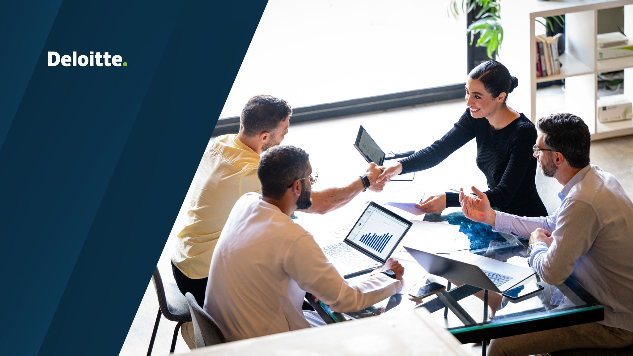 Business professionals shaking hands and discussing work around a meeting table with laptops and documents in a modern office setting.