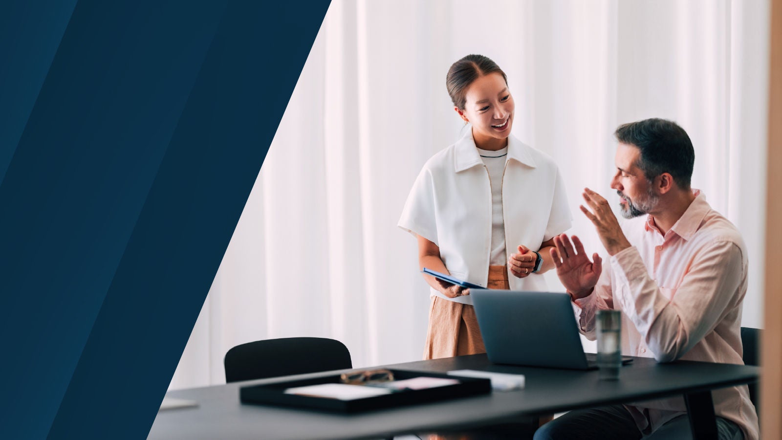 Two professionals collaborating in an office. A woman stands smiling while holding a tablet, as a seated man gestures while speaking. A laptop and a water glass are on the desk between them.