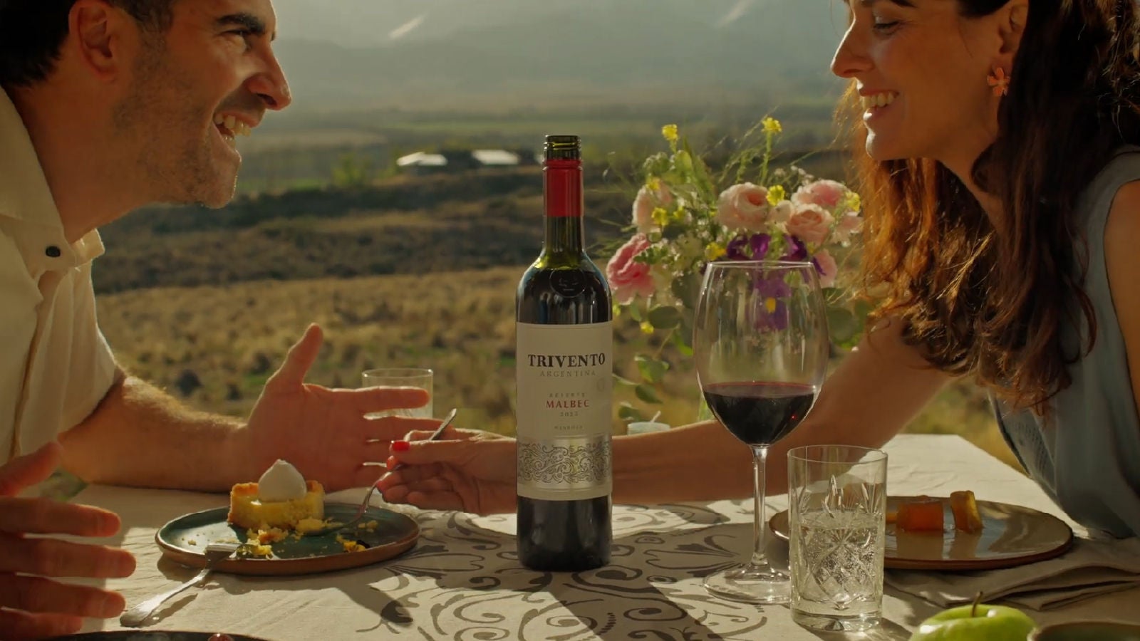 Man and woman laughing and drinking wine at a table overlooking a vineyard