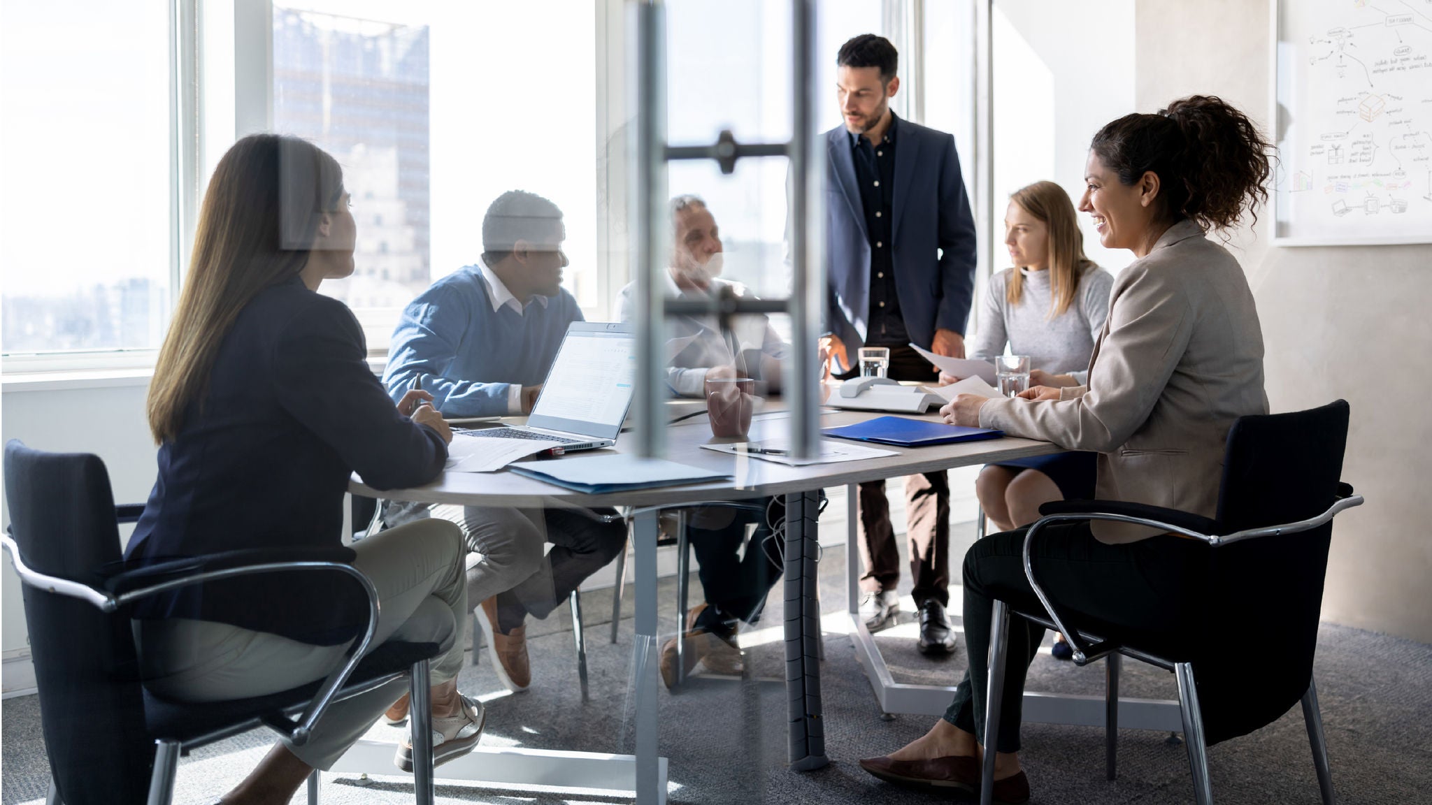 A group of diverse professionals engaged in a meeting around a glass-walled conference table, with a whiteboard full of handwritten notes in the background.