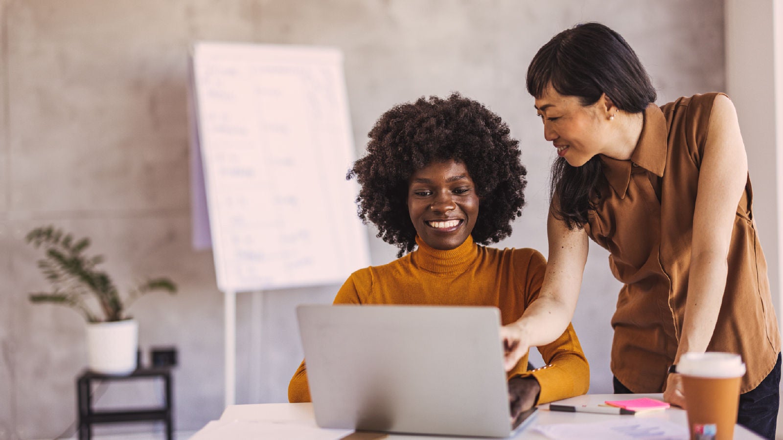 Two women smiling and working together on a laptop in a casual office setting.