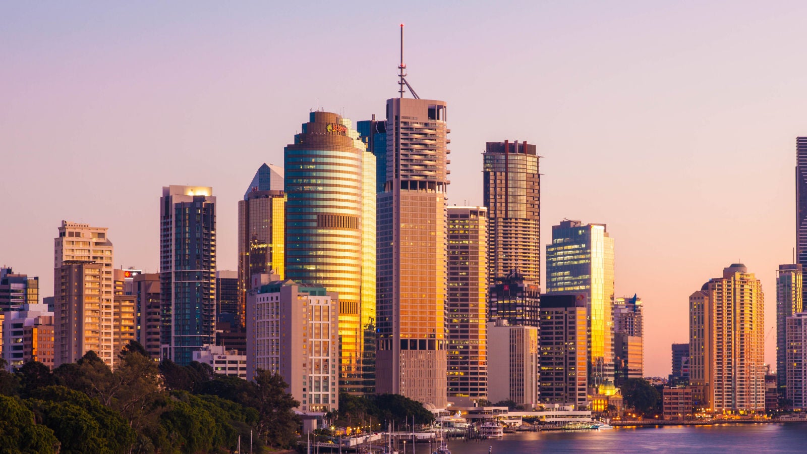 Brisbane city skyline at sunset, with golden reflections on high-rise buildings and sailboats docked along the riverfront.