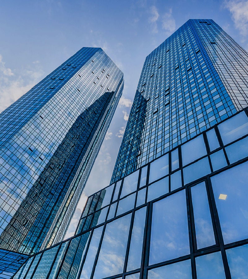 Upward view of two modern glass skyscrapers reflecting the blue sky and clouds.