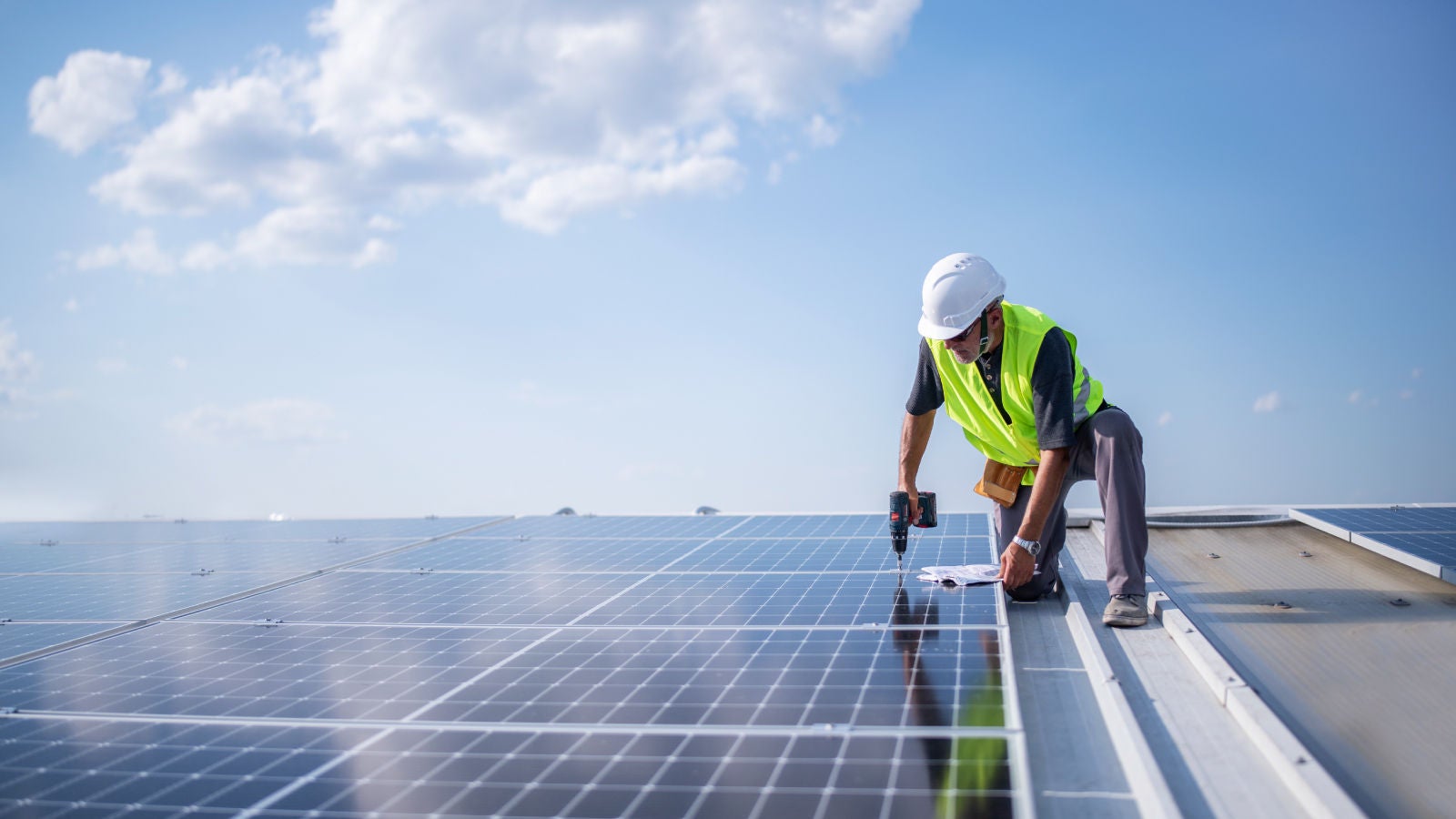 Technician in a safety vest and hard hat working on rooftop solar panels under a bright blue sky with scattered clouds.
