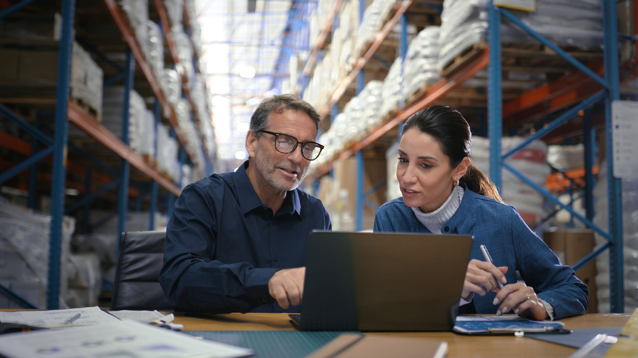 Man and woman seated at a desk in a warehouse, reviewing information on a laptop together.