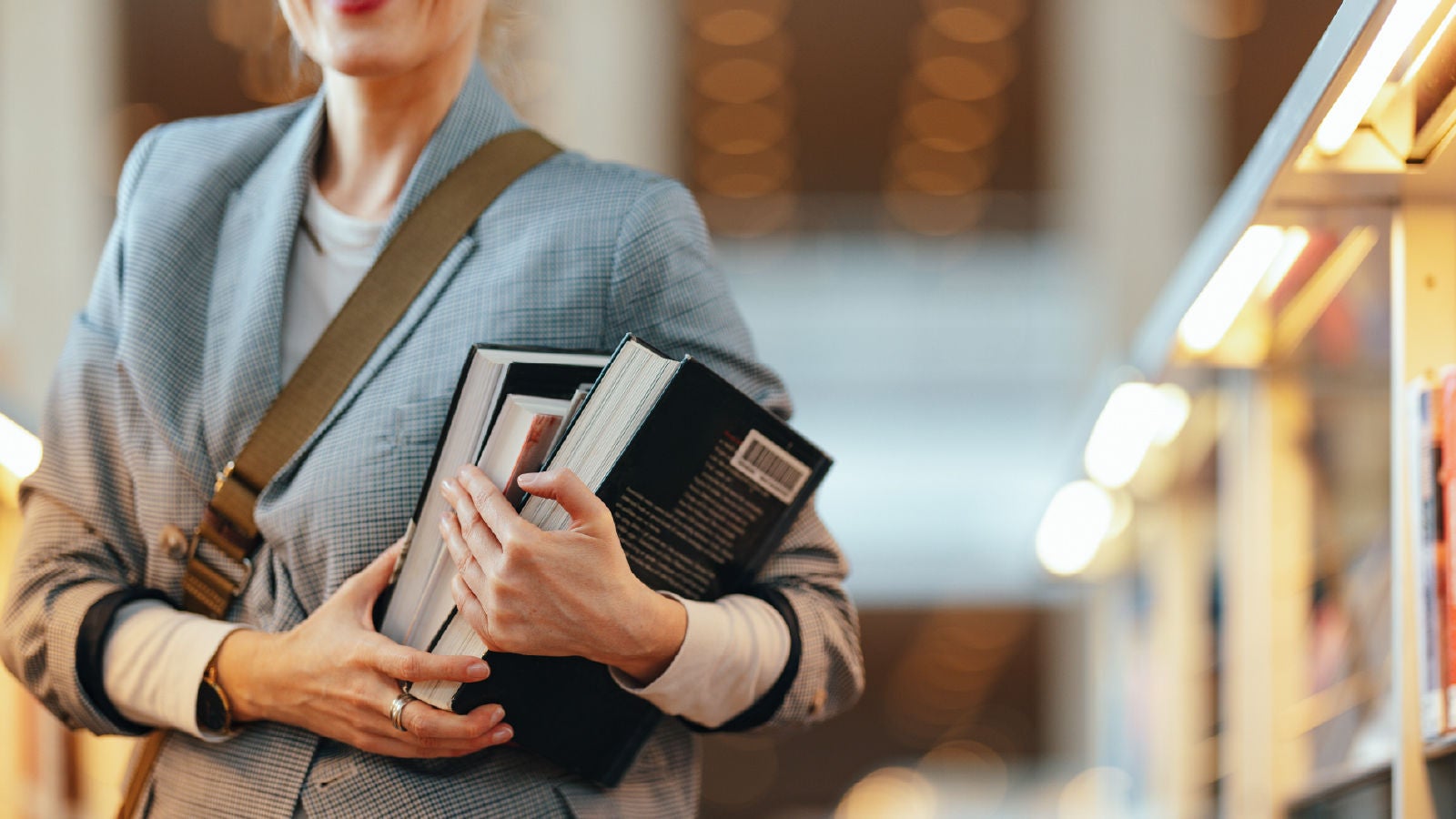 A woman in a plaid blazer holding several books close to her chest, standing between bookshelves in a brightly lit library.