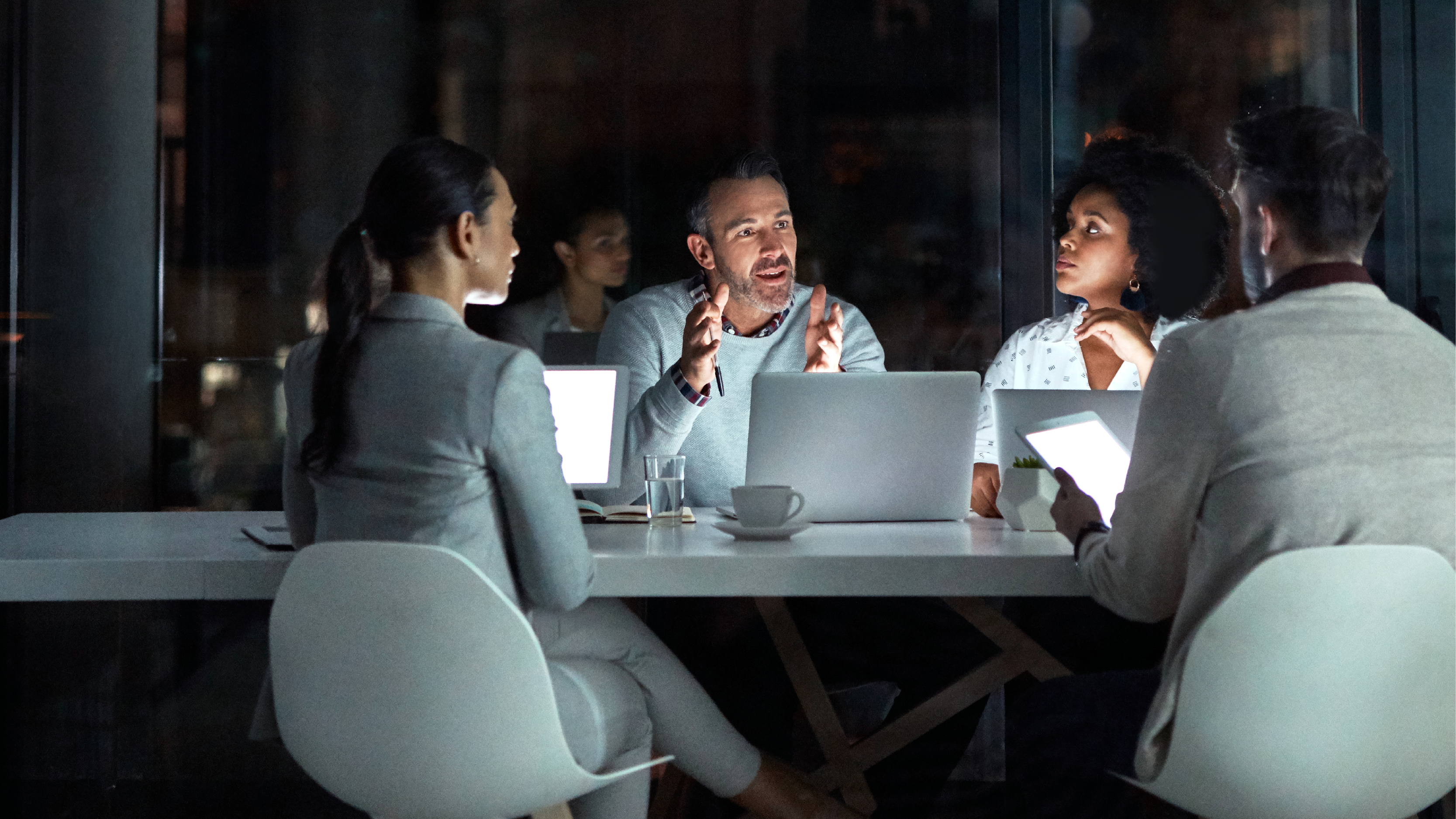 A group of colleagues gathered at a table during a nighttime meeting, illuminated by screens as they discuss and collaborate.