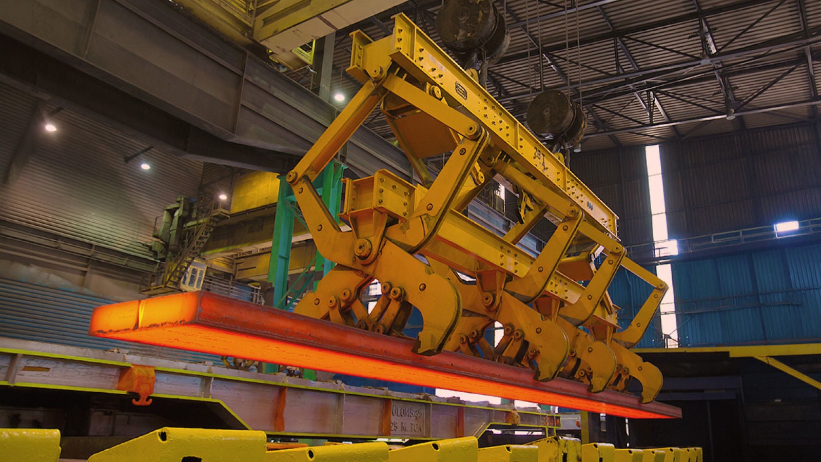 Industrial machinery lifting a glowing hot steel slab inside a manufacturing facility.