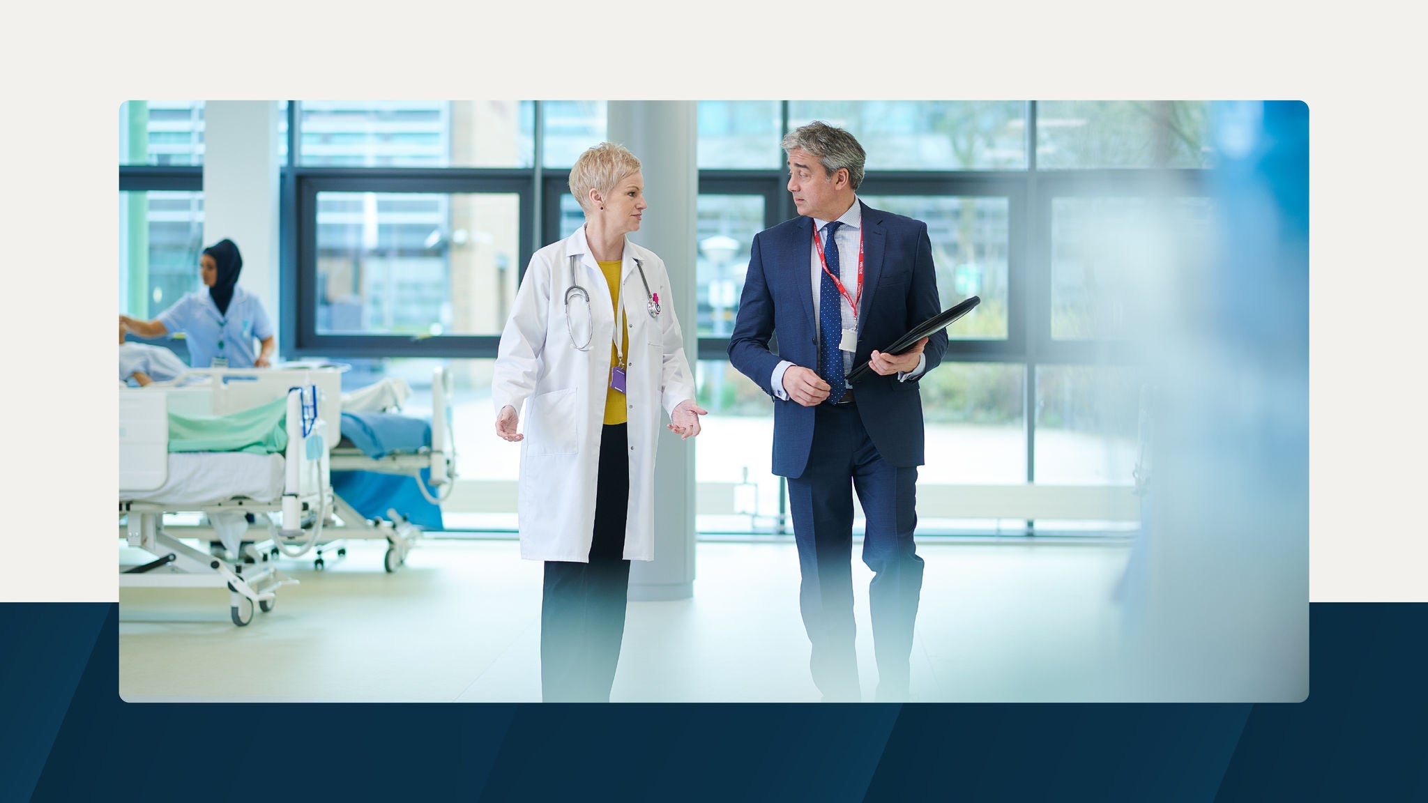 A doctor and a suited professional having a discussion while walking through a hospital corridor, with medical staff and patient beds visible in the background.