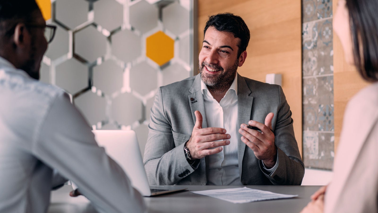 Business professional in a gray suit speaking and gesturing while seated at a table with two colleagues, with a laptop and documents visible.
