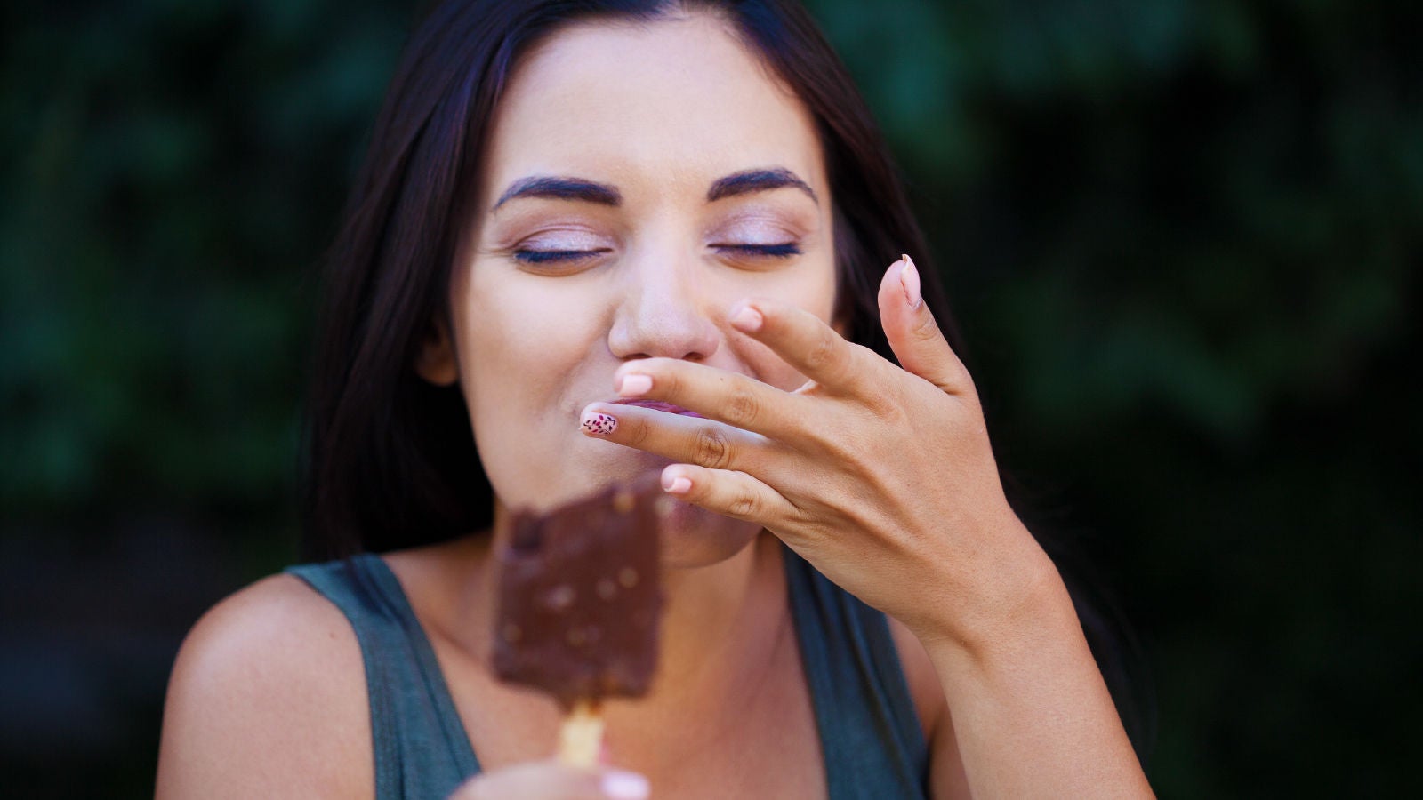 Close-up of a woman enjoying a chocolate ice cream bar, smiling with her eyes closed and touching her lips, symbolizing satisfaction and indulgence.