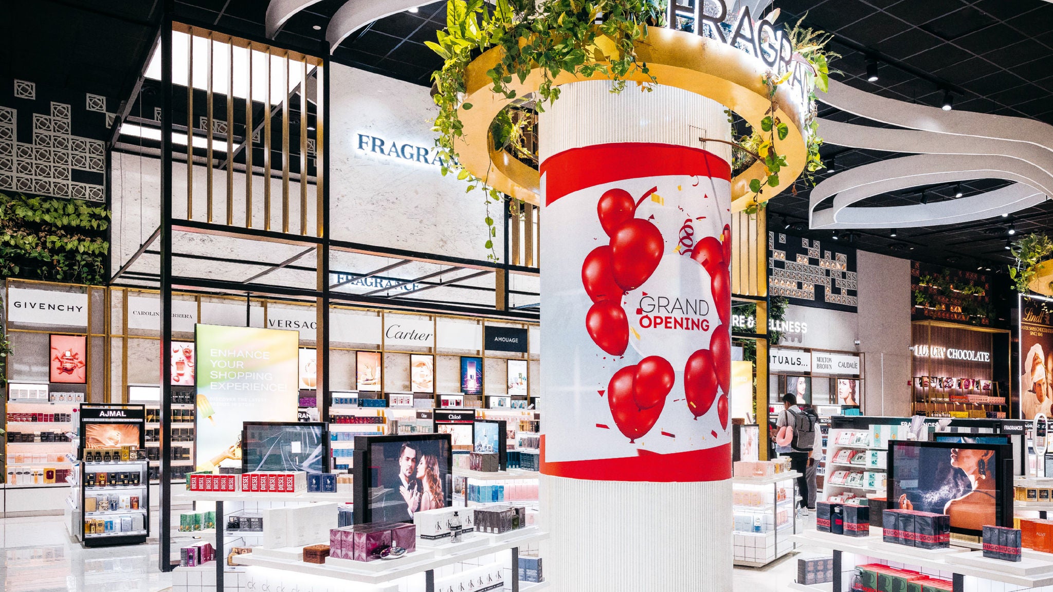 A brightly lit retail store interior with fragrance displays, brand signage, and a large cylindrical column featuring a “Grand Opening” sign with red balloons.