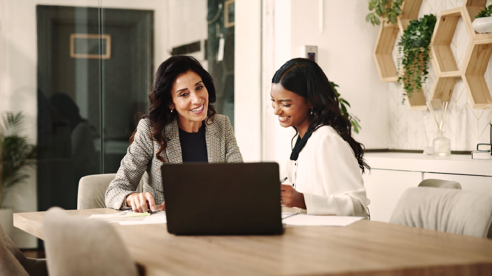 Two women in business attire sit at a desk with a laptop, smiling and discussing work in a modern office space with plants and shelving in the background.