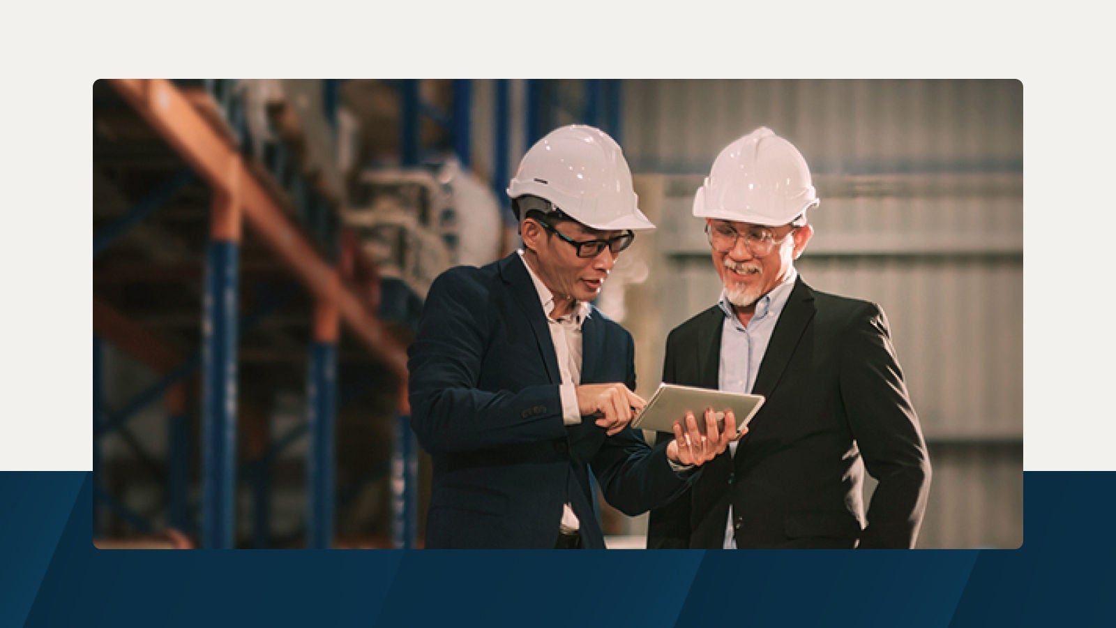 Two professionals in business attire and hard hats discuss production plans on a tablet in a warehouse setting.