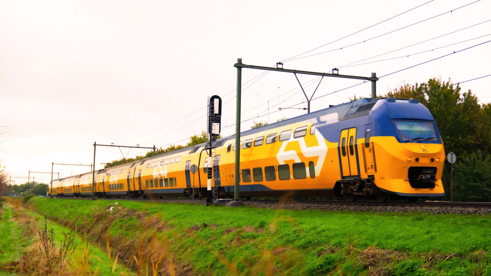 Yellow double-decker passenger train traveling along electrified tracks through green countryside.