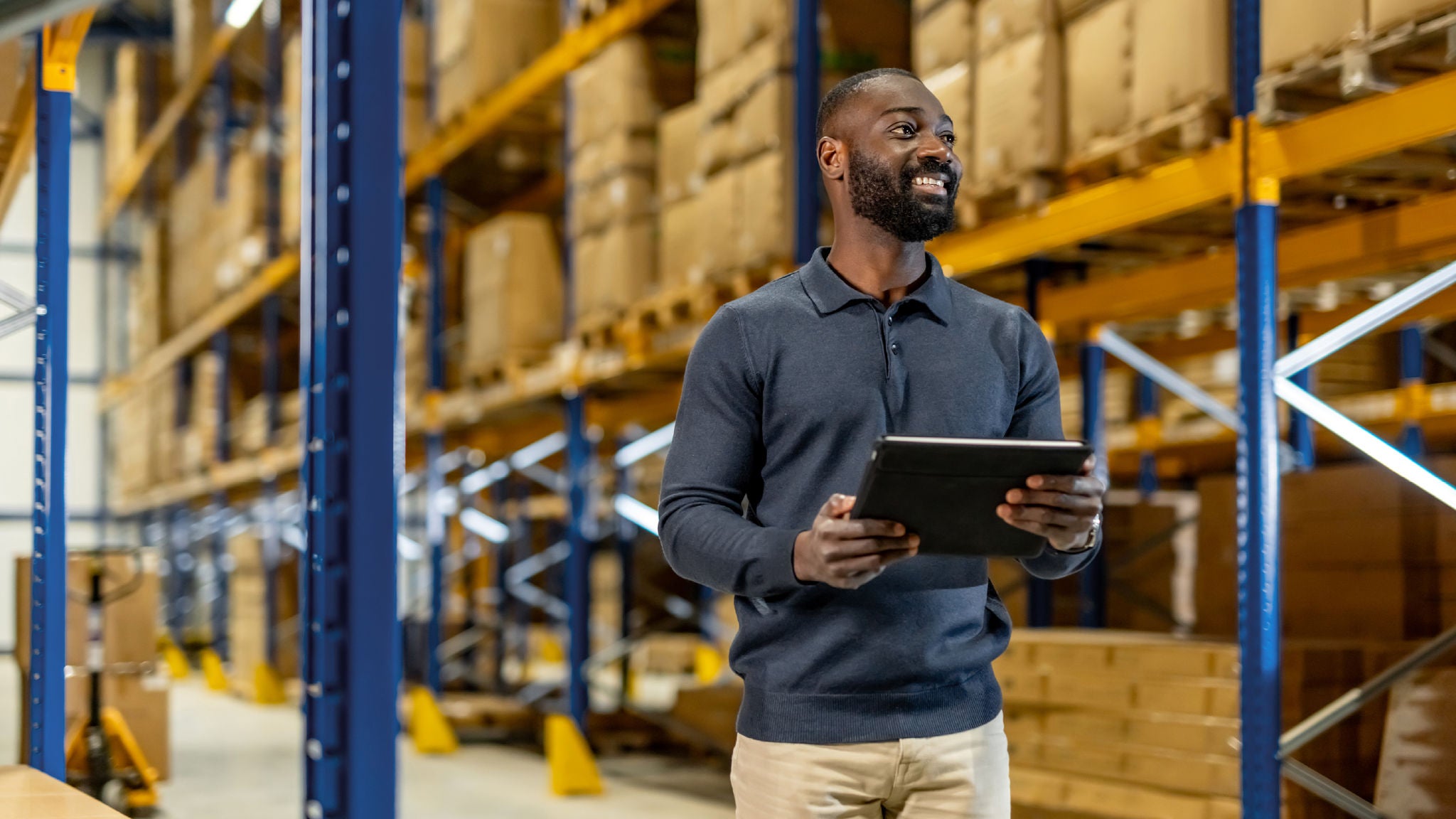 A man standing in a warehouse holding a tablet, with tall shelves of boxes in the background.