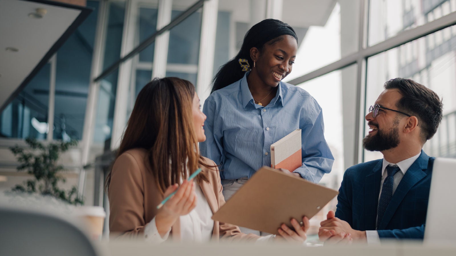 Three colleagues in a modern office meet around a table, reviewing documents and a laptop. One woman stands holding notebooks while talking with a seated woman and a man in a suit who is smiling as they work together.