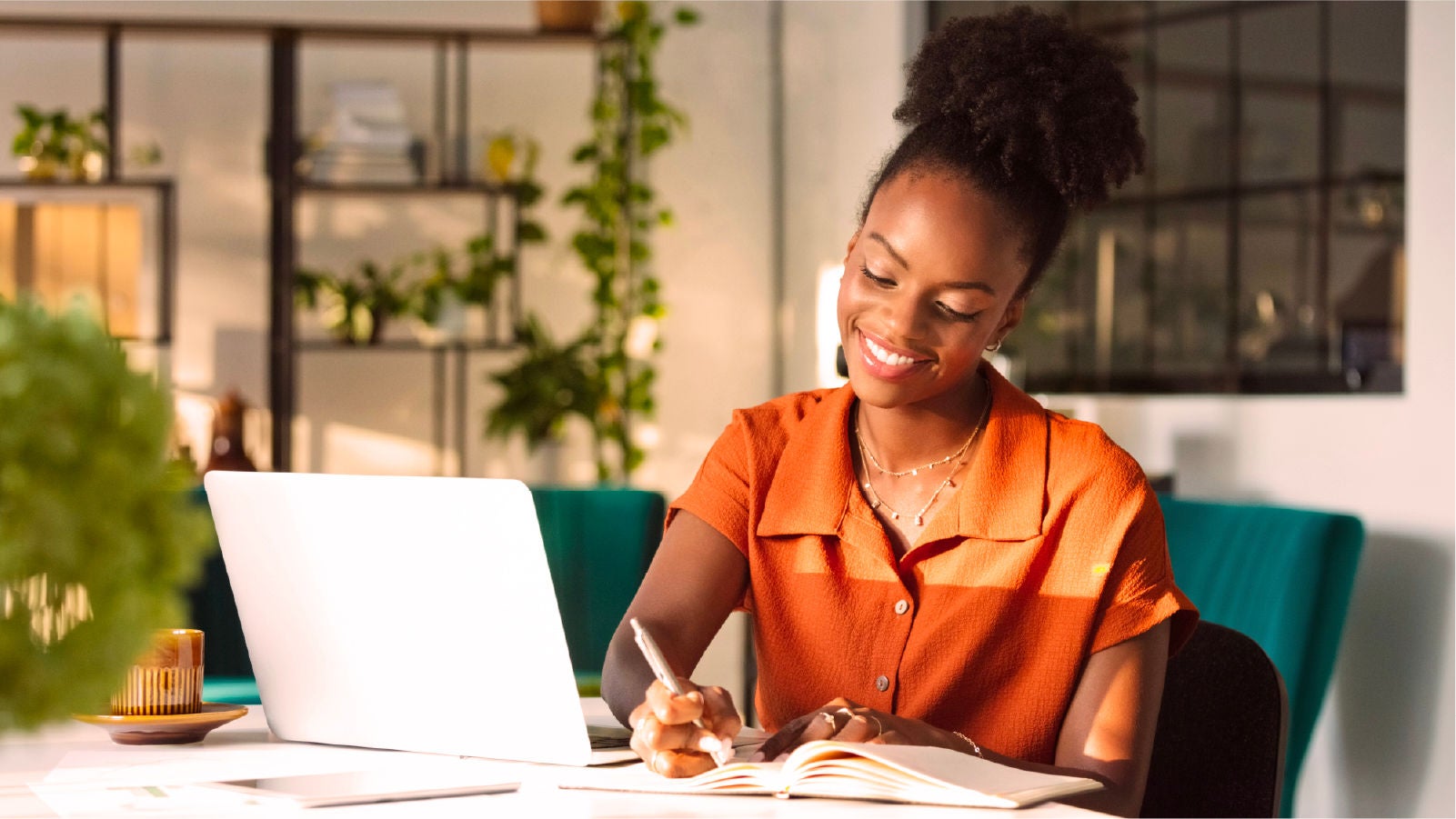 A woman smiling while writing in a notebook at a desk with a laptop in a well-lit office space.