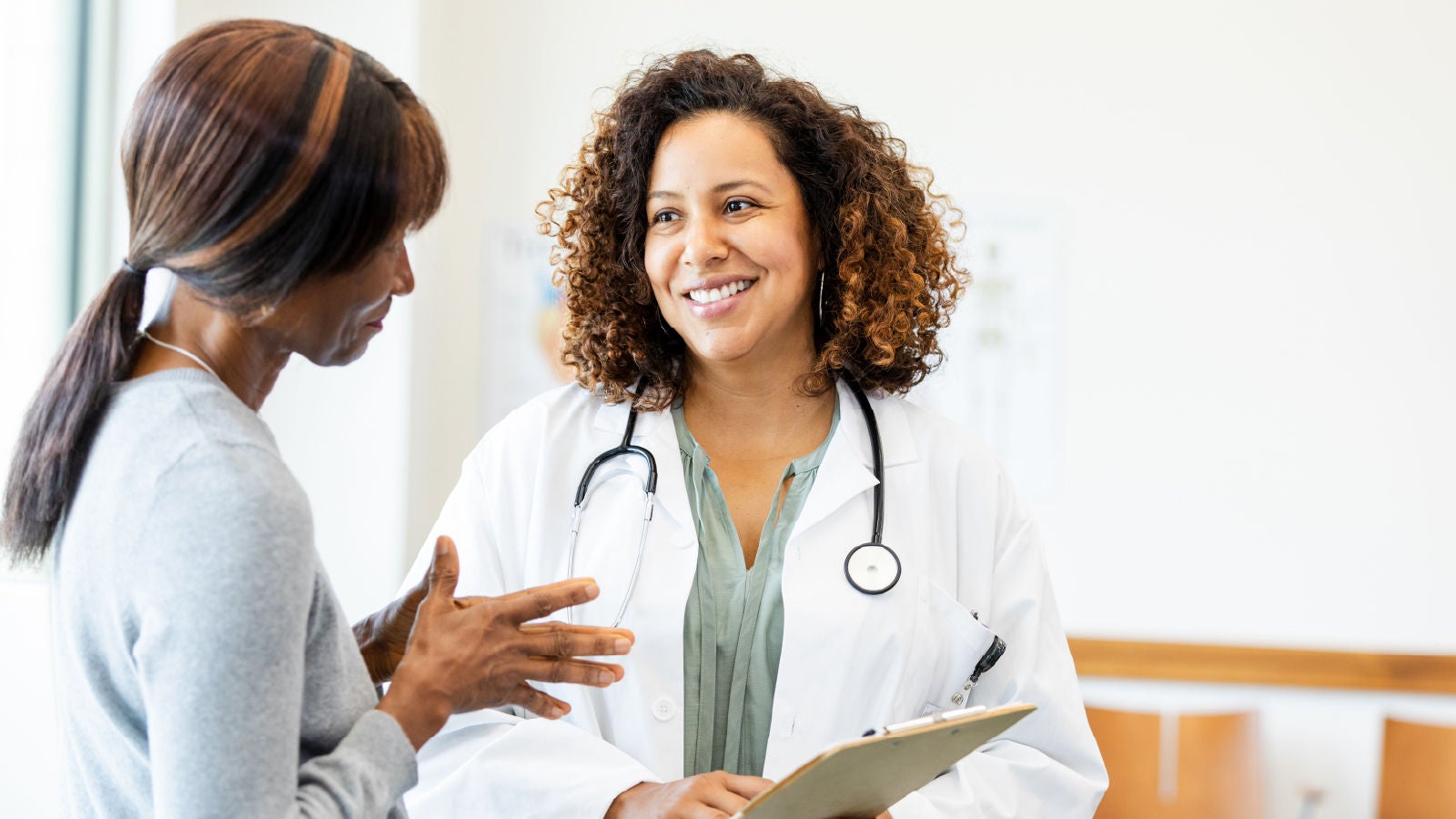 Image showing a doctor in a white coat with a stethoscope smiling while speaking with a patient holding a clipboard.