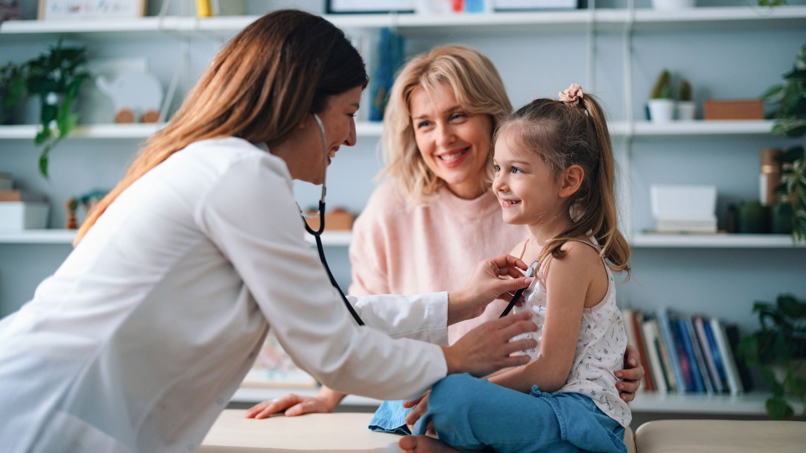 Healthcare professional examining a smiling young girl with a stethoscope as her mother watches.
