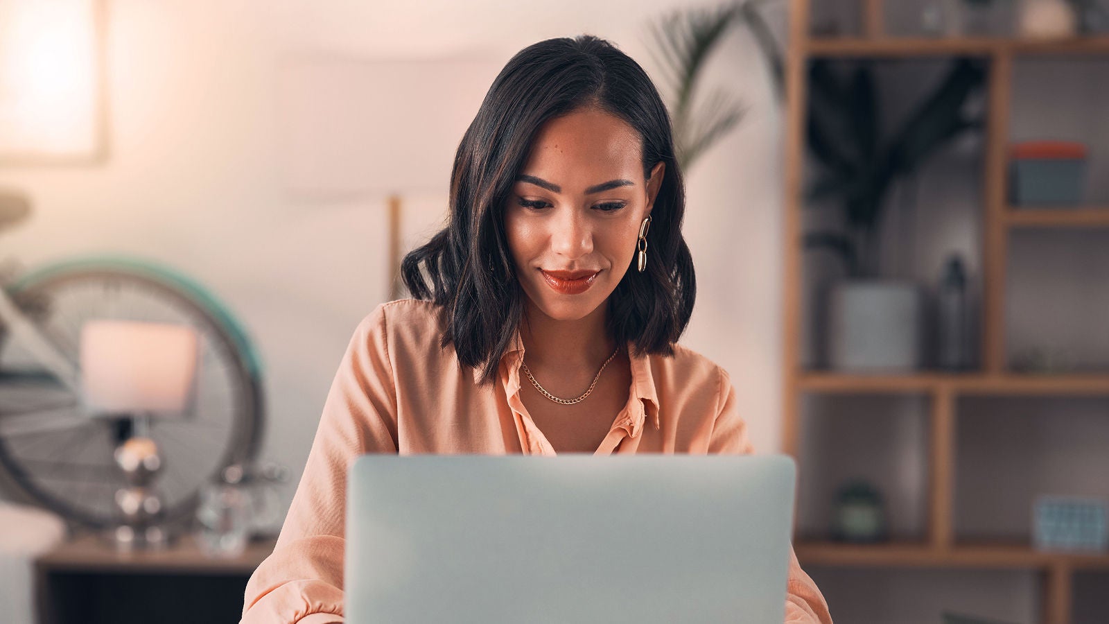A professional woman with shoulder-length dark hair, wearing a peach blouse, works on a laptop in a well-organized home office, symbolizing strategic planning and productivity.