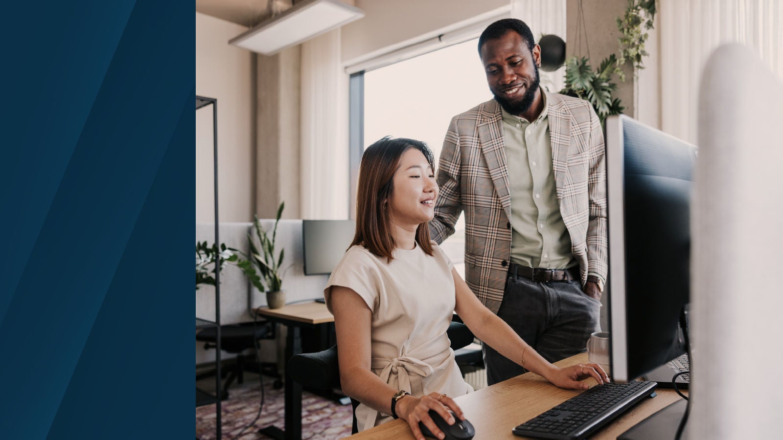 Two professionals collaborate at a desk in a modern office, with one seated using a desktop computer and another standing beside her, while plants and workstations are visible in the background.
