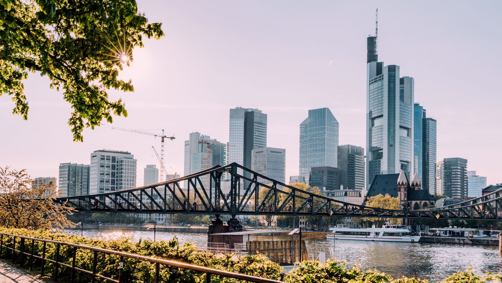 A city skyline with modern buildings, a river, and a bridge with people walking across, surrounded by greenery and trees.
