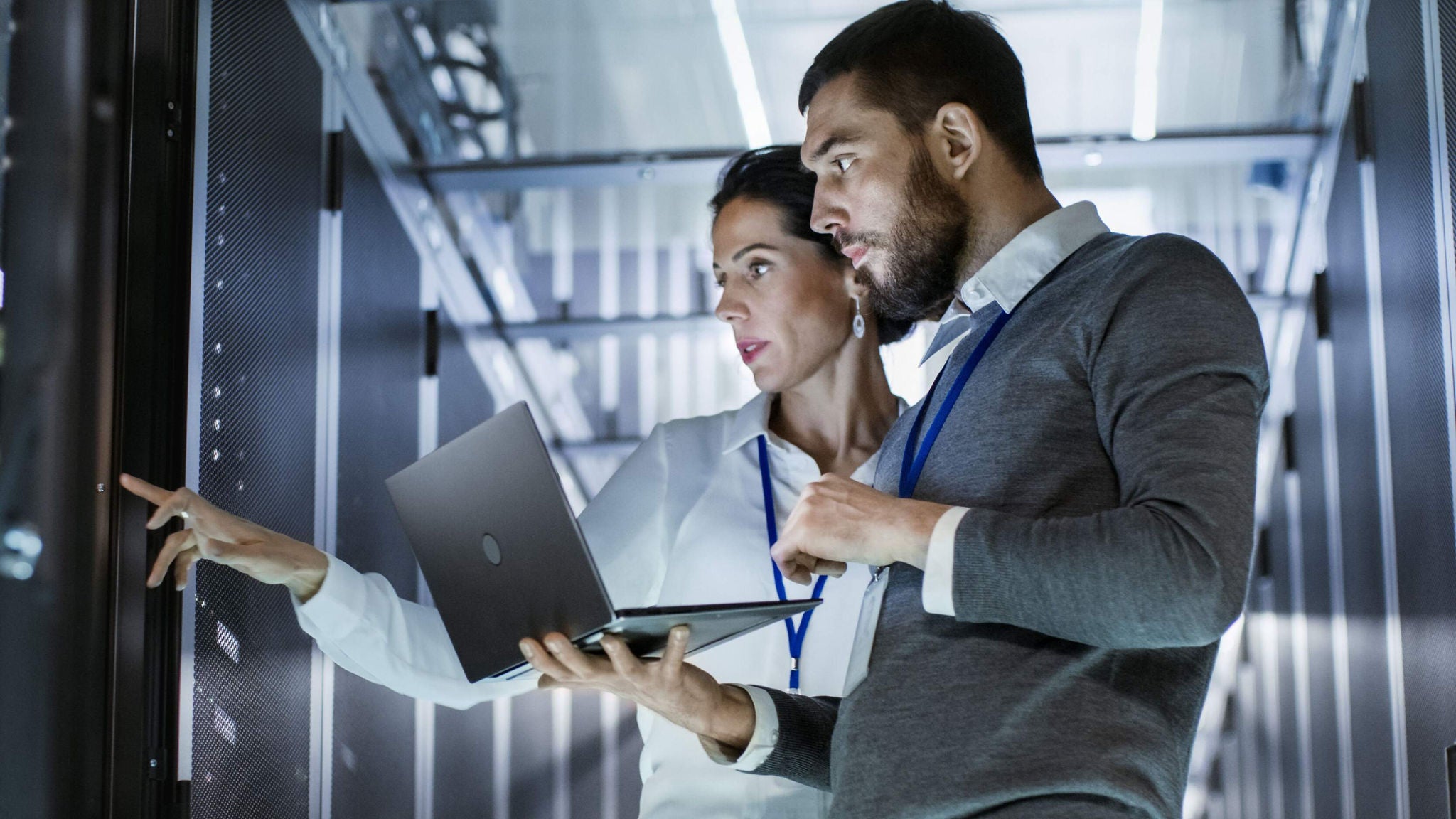 Two IT professionals inspect a server rack, with one holding a laptop and the other pointing at equipment.