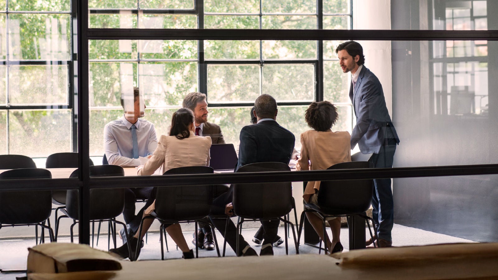 A wider view of a team of professionals meeting in a glass-walled conference room, with participants seated and discussing work materials.