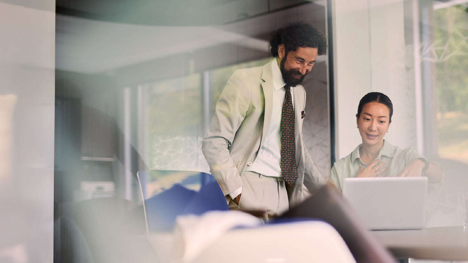 A man in a suit and a woman collaborating at a laptop in a bright glass-walled office, seen through a reflective surface.