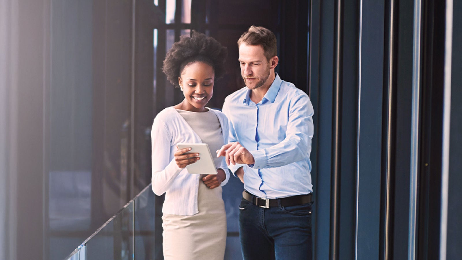 Two professionals standing in a modern office hallway, reviewing information on a tablet together.