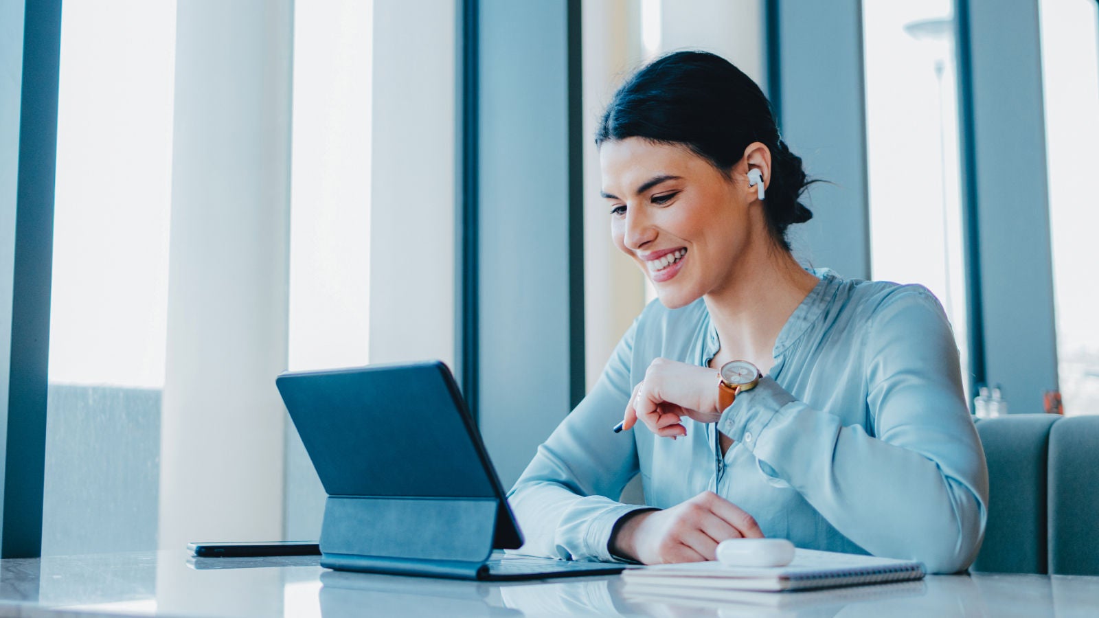 Professional wearing wireless earbuds and smiling while working on a tablet at a desk.
