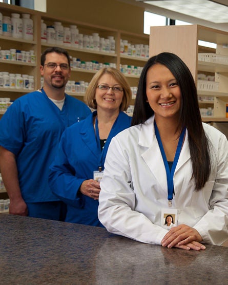 Three healthcare professionals stand in a pharmacy. A woman in a white lab coat smiles at the counter in front, with two colleagues in blue scrubs standing behind her. Shelves filled with medication bottles line the background.