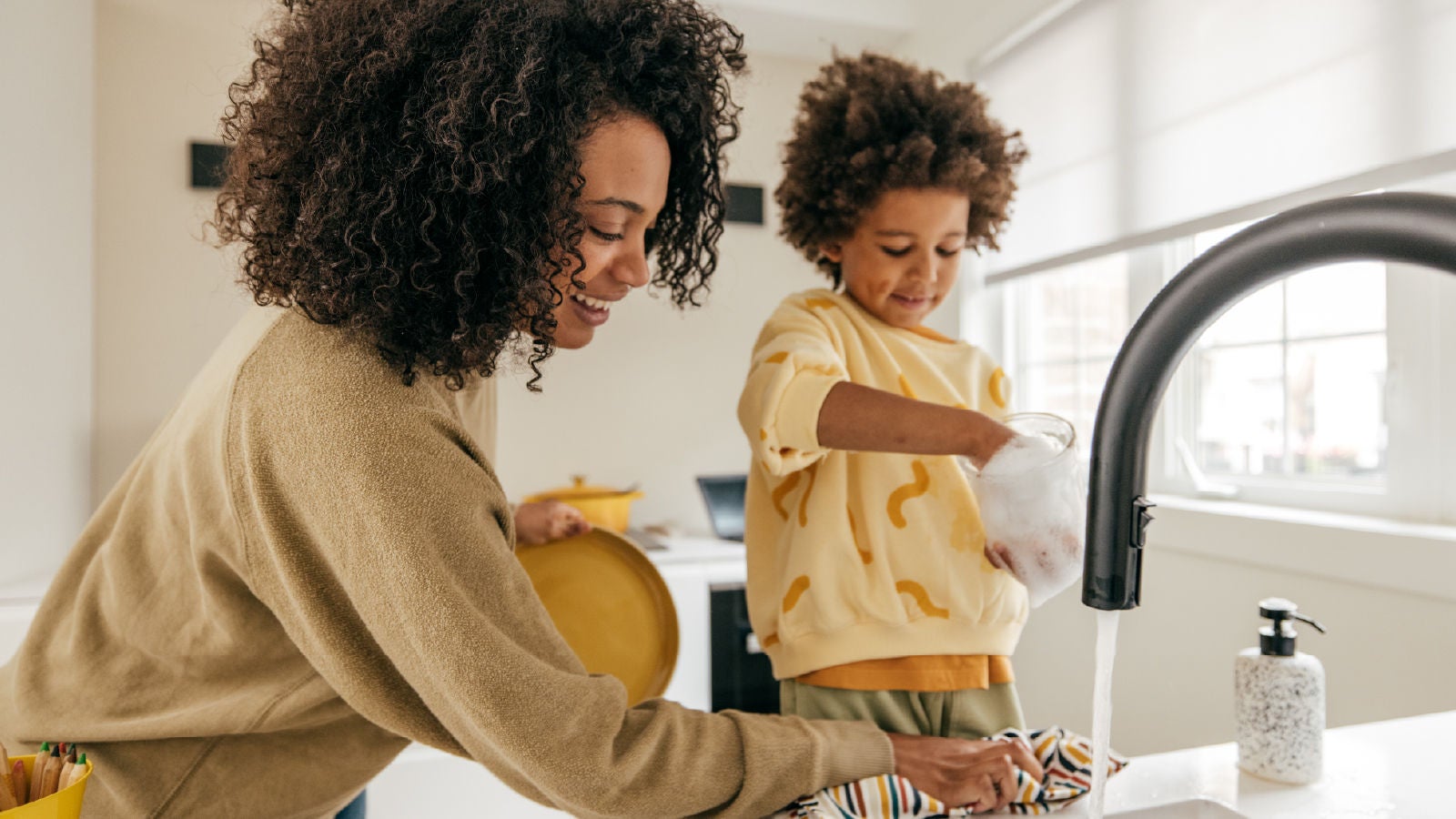 A woman and child washing dishes together in a bright kitchen, smiling and enjoying the task, representing family connection and everyday moments.