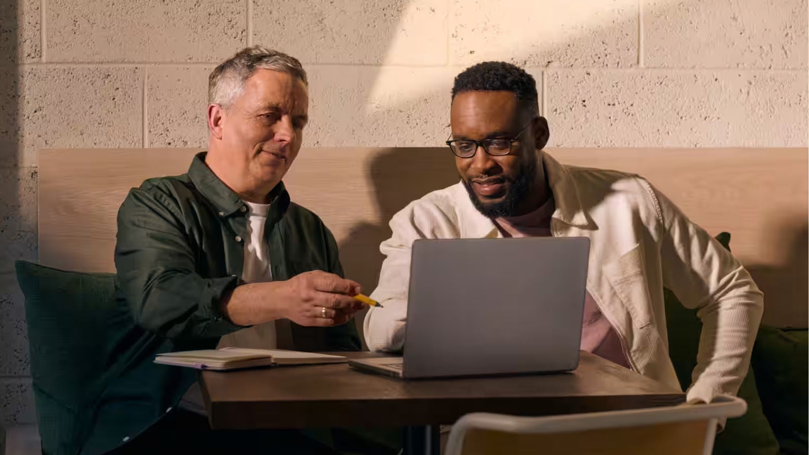 Two colleagues seated at a table reviewing information together on a laptop in a meeting space.
