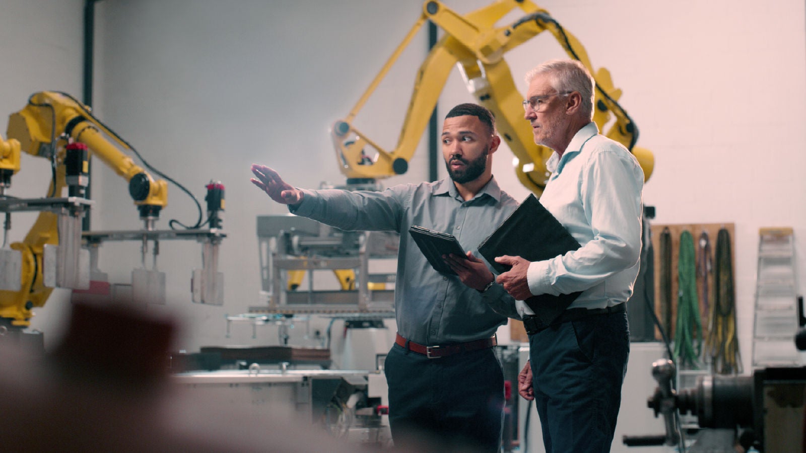 Two men in a factory setting with large yellow robotic arms. One man is holding a tablet and gesturing toward the machinery, while the other, holding a folder, listens attentively.