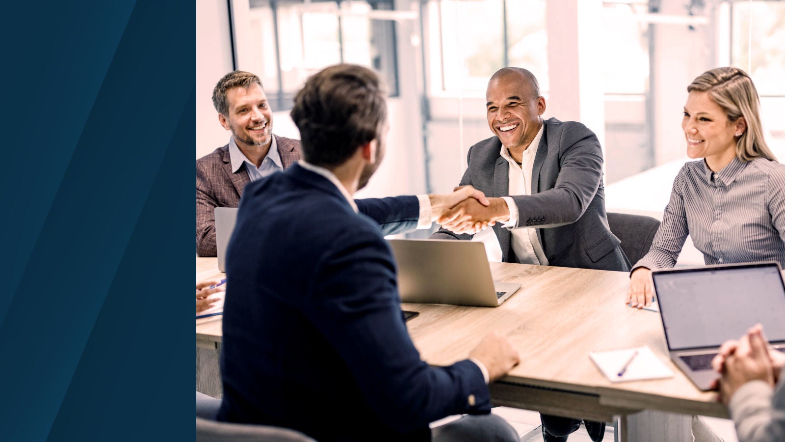Four professionals in a meeting room smile as two men shake hands across a table with laptops and notebooks.