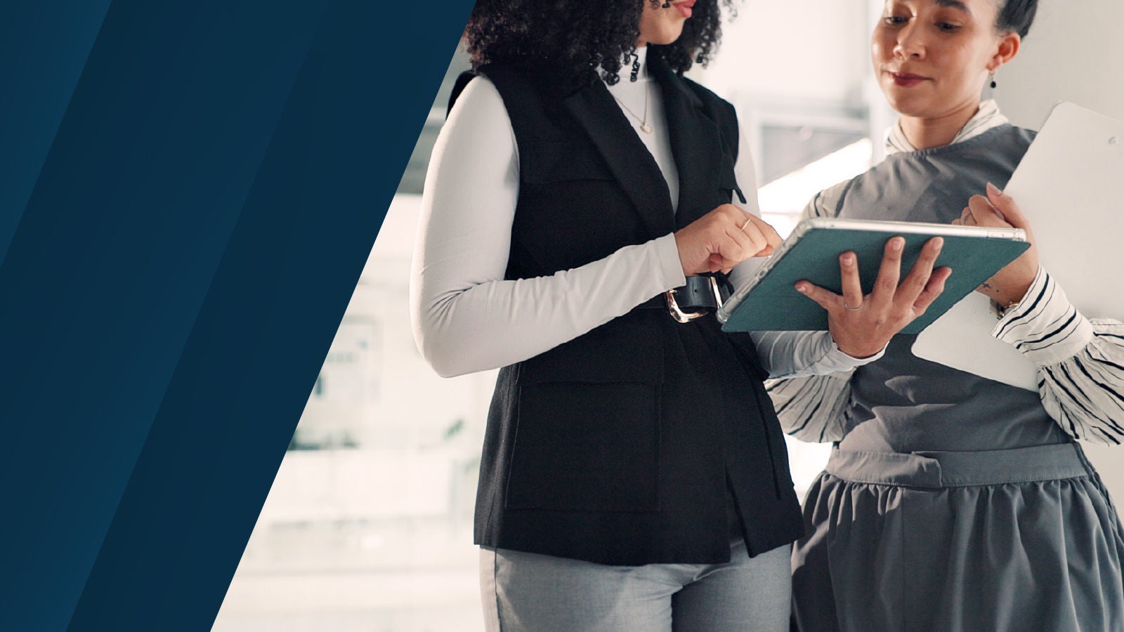 Two professionals standing together, one holding a tablet and the other pointing at it. The left side has a dark blue diagonal overlay