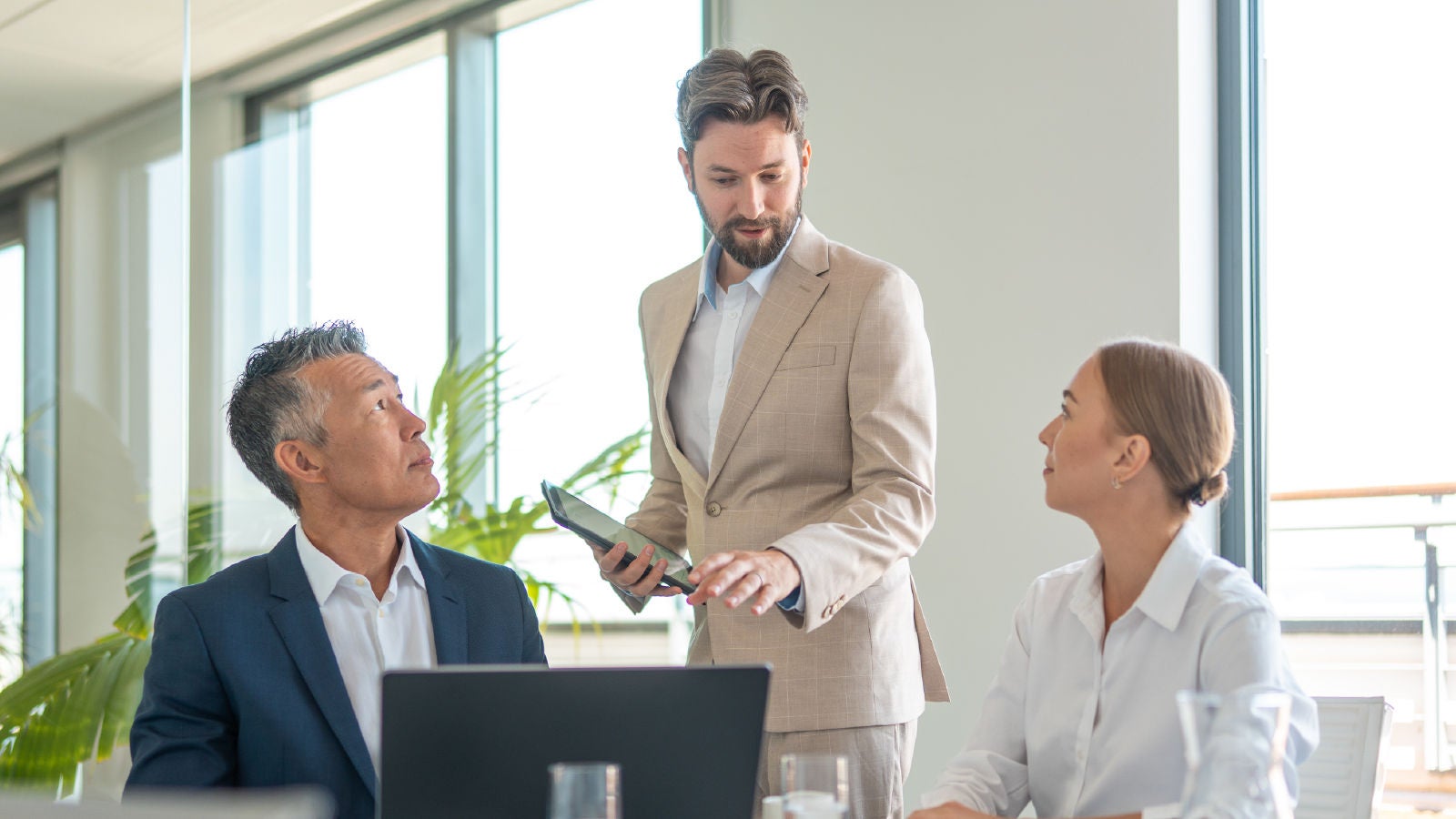 A man in a beige suit stands holding a tablet and speaks to two seated colleagues—one older man in a navy suit and one woman in a white blouse—during a meeting in a bright office with greenery in the background.