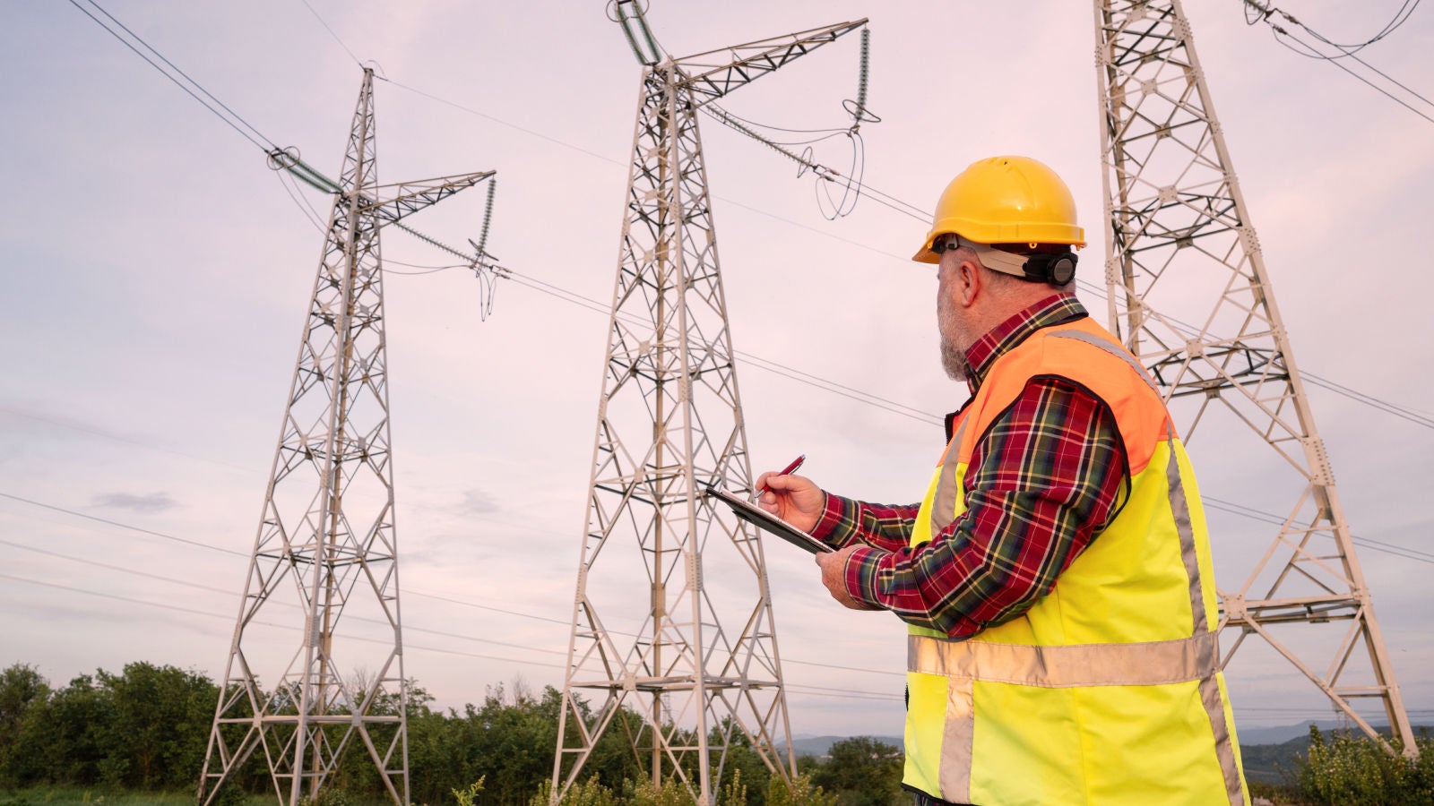 Utility worker in a safety vest and hard hat inspecting electrical transmission towers while holding a clipboard.