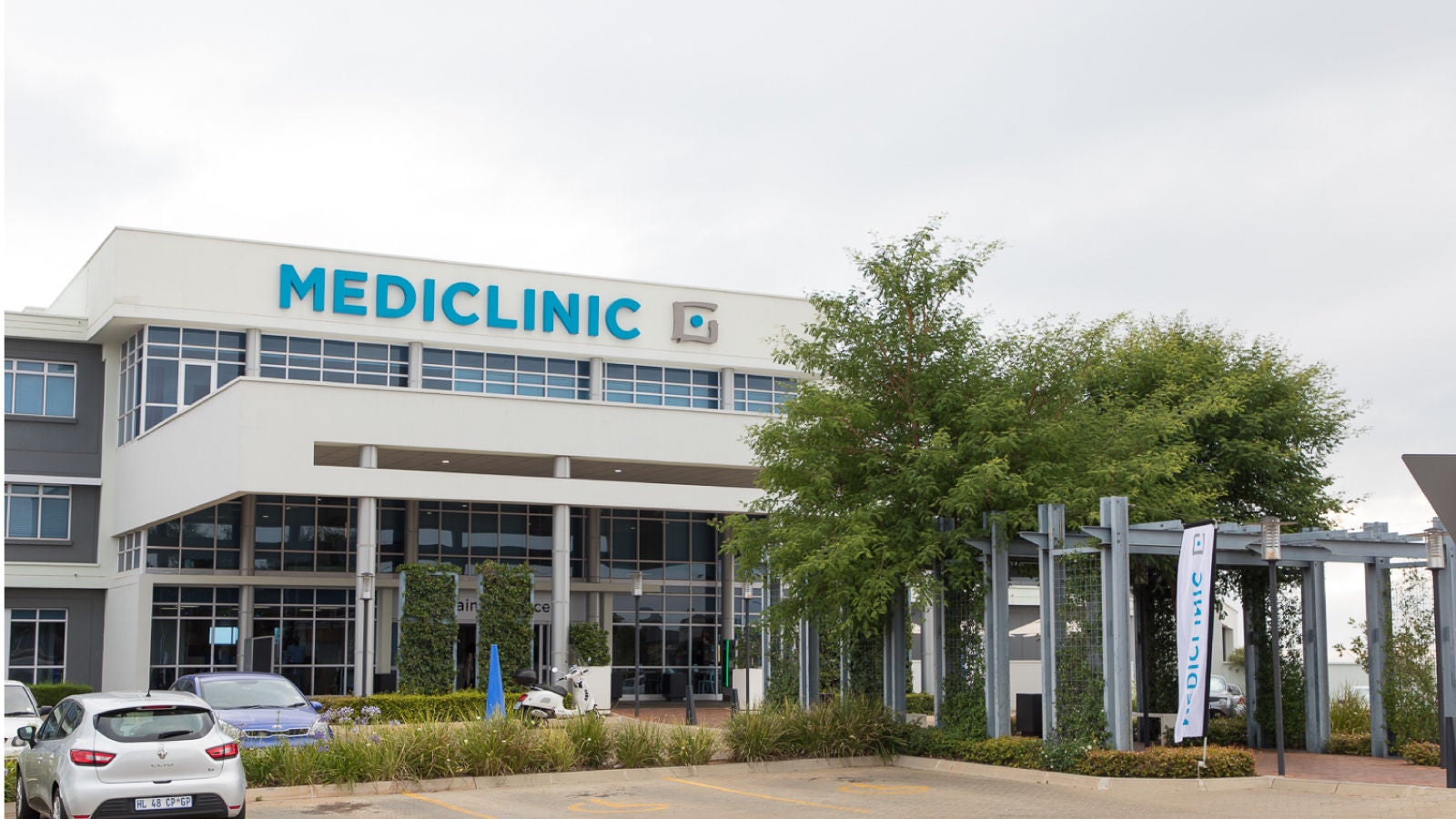 The exterior of a Mediclinic facility, featuring large glass windows, a prominent "Mediclinic" sign, and a landscaped entrance with greenery and parked vehicles.