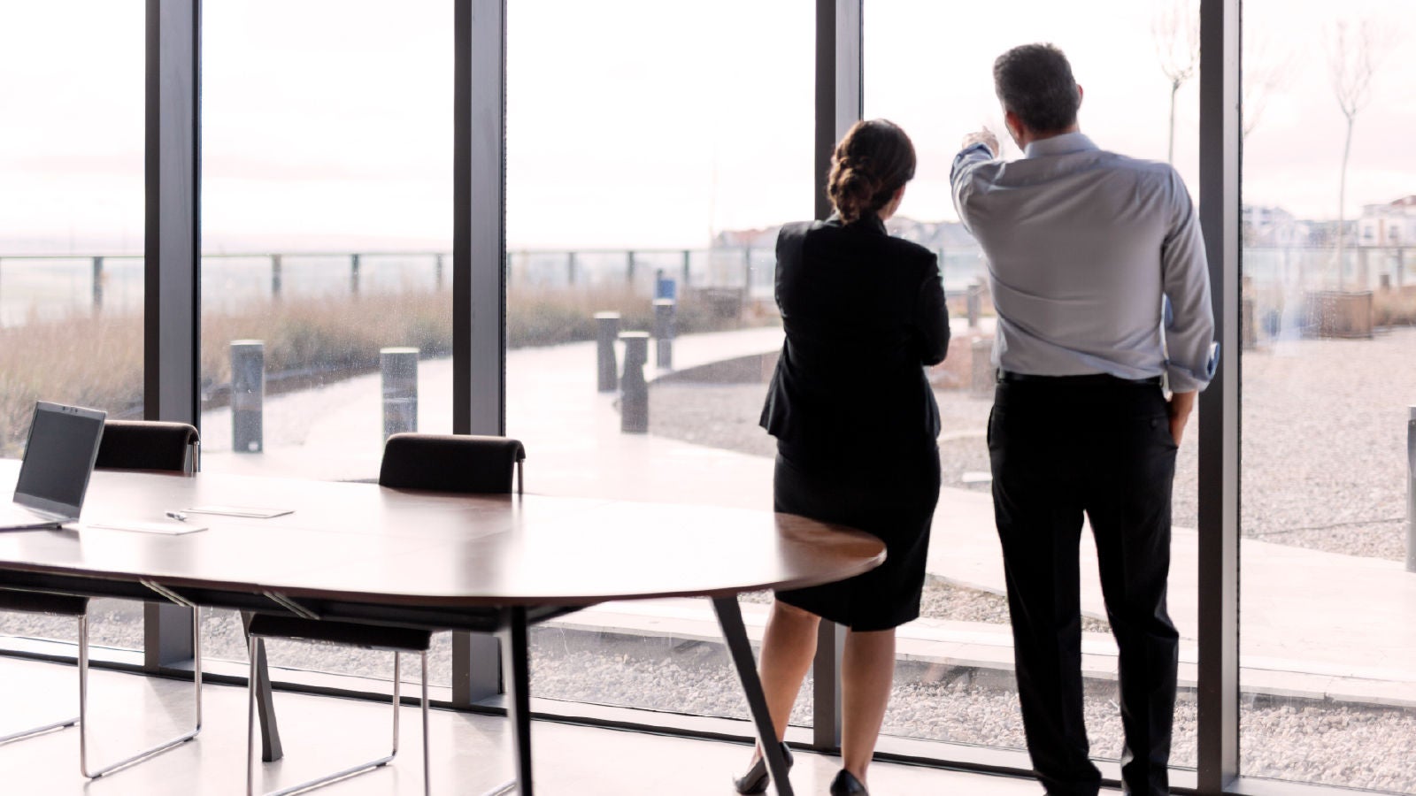 Two executives standing near a floor-to-ceiling window in an office, looking outside while having a discussion. A conference table, chairs, and a laptop are visible in the foreground.