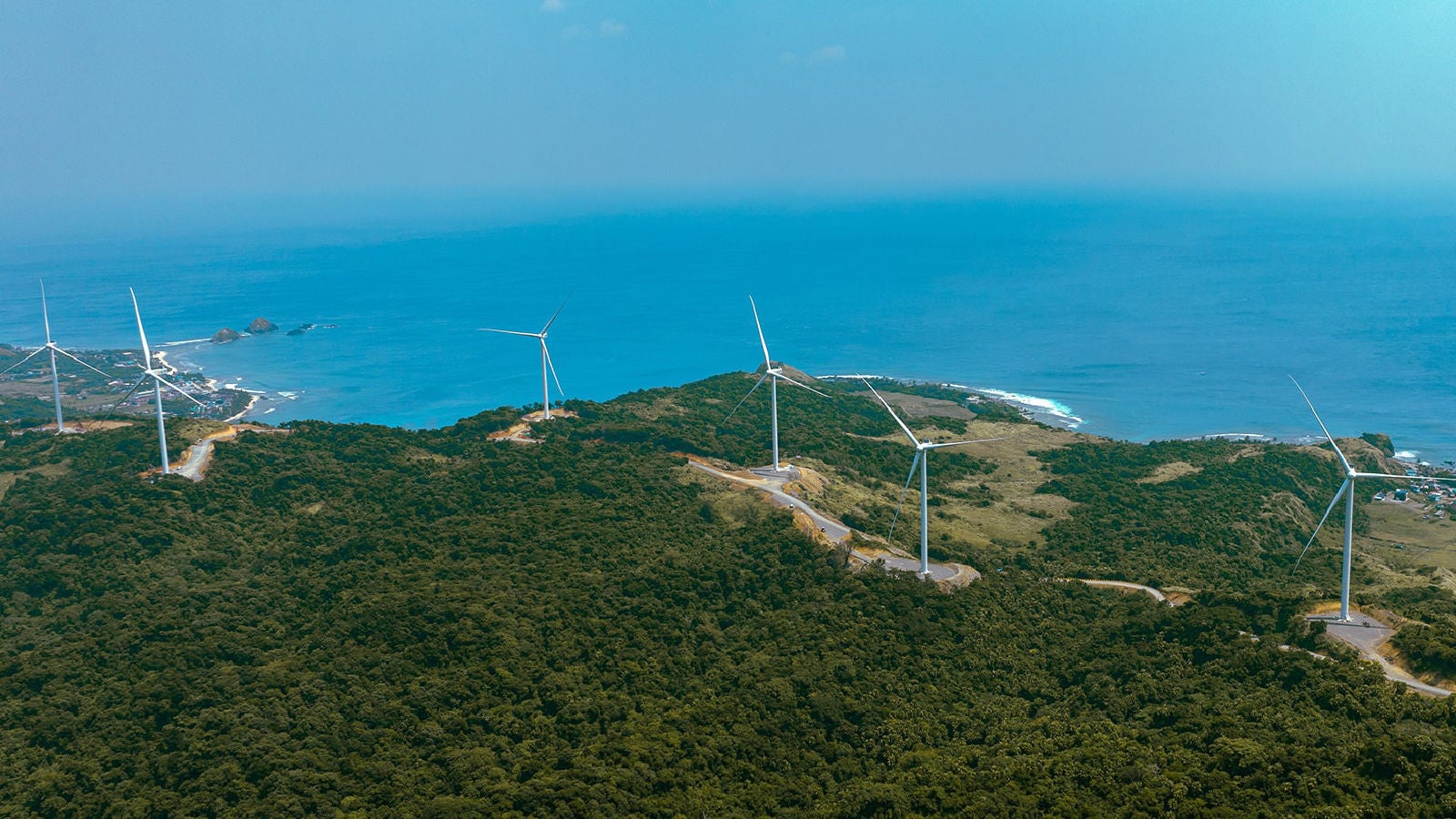 Wind turbines installed along a coastal hillside overlooking the ocean under a clear blue sky.