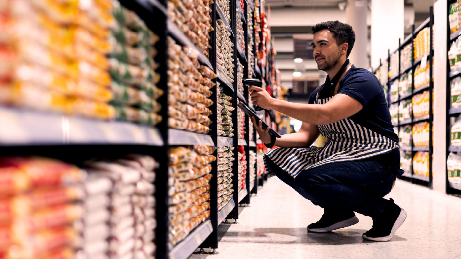 A grocery store worker in a striped apron crouches in an aisle, scanning products with a barcode reader and checking a tablet, surrounded by shelves stocked with goods.