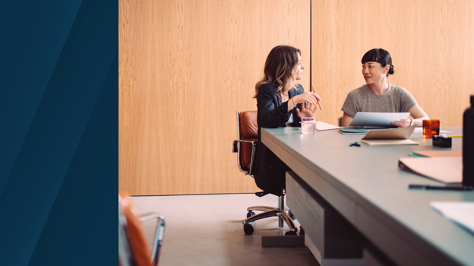 Two professionals having a discussion at a meeting table with papers and laptops, set against a light wood-paneled wall in an office.