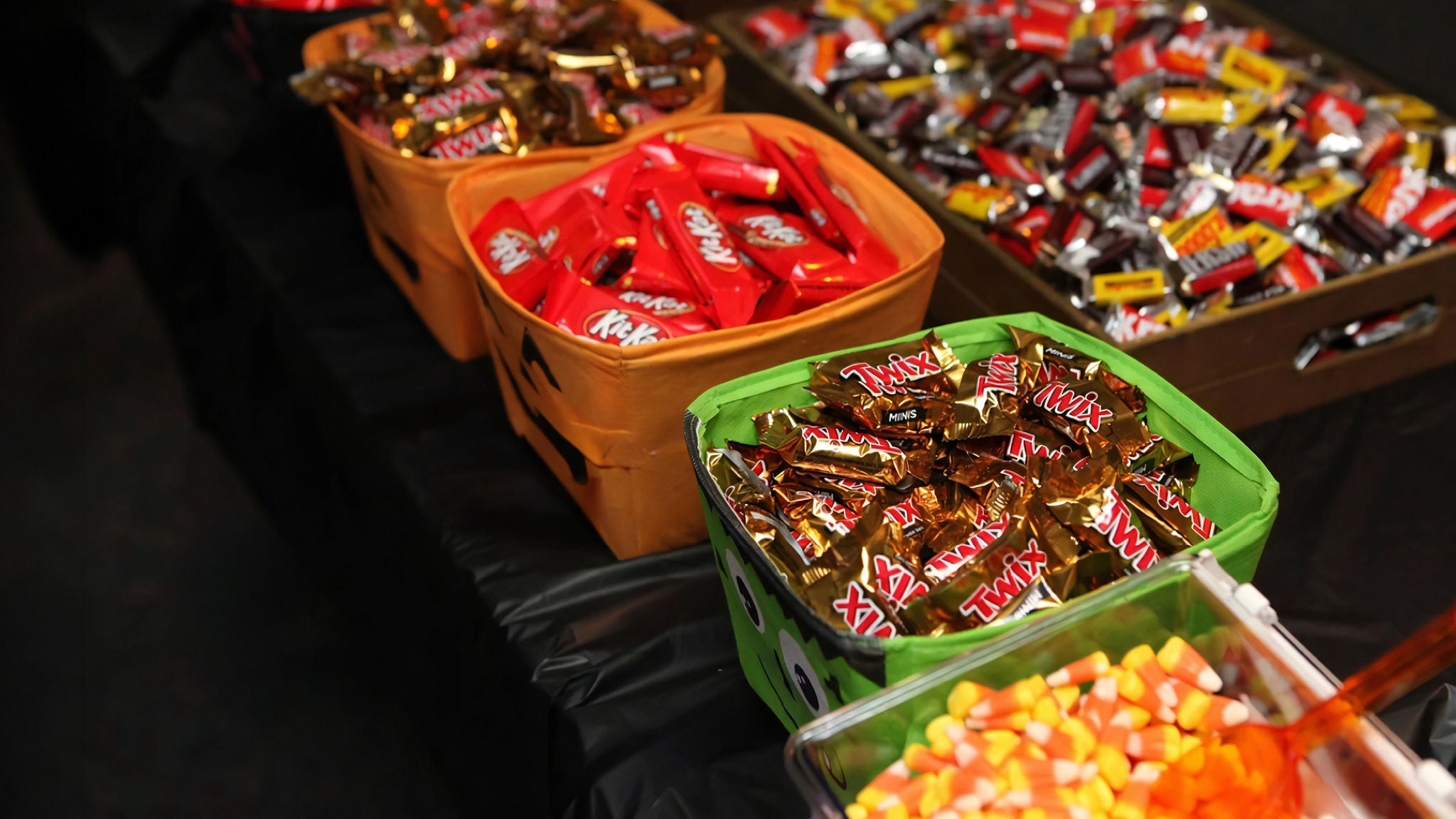 Wide shot of colorful Halloween candy display with bins filled with Twix, KitKat, and other chocolate bars, along with candy corn in the foreground.