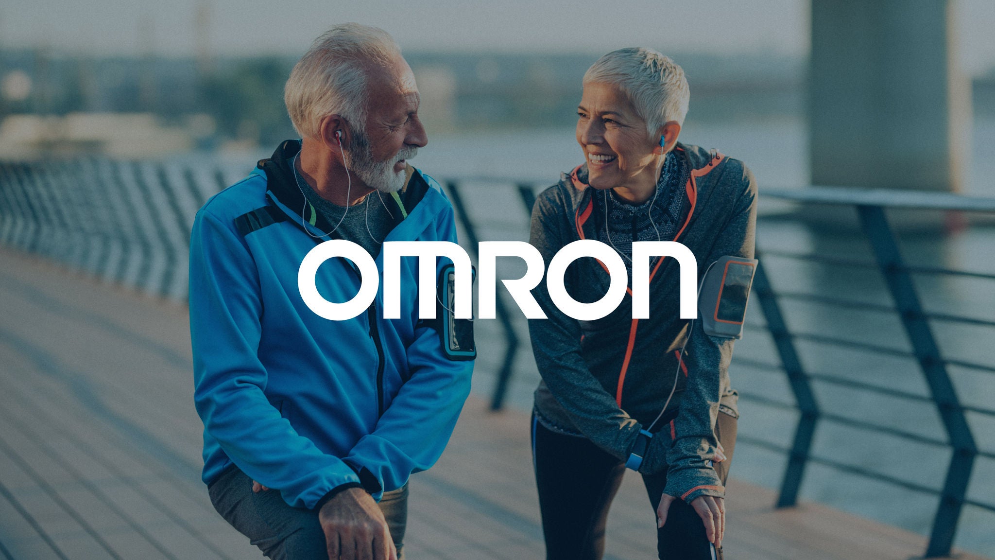 Omron logo displayed over a dimmed background of two older adults smiling together after exercising by the water.